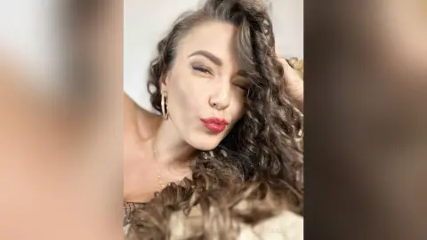 MontseFerrera winks while running her hand through her long, wavy brown hair and wearing red lipstick.