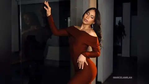 MarcelaMilan poses in a tight, off-the-shoulder brown dress with her eyes closed and long dark hair flowing.