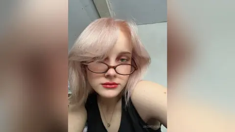 Young woman with pink hair and glasses wearing a black tank top in a soft, indoor setting.