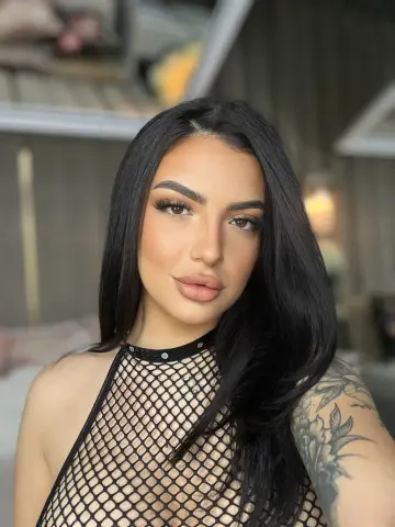 NoraSin poses with long black hair, wearing a black fishnet top and a black choker, showing off a large floral tattoo on her shoulder.