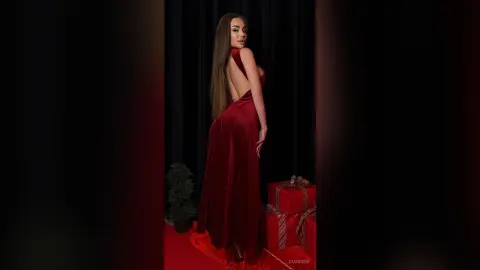 Elegant woman in a long red velvet gown posing next to Christmas gifts in a moody festive setting.
