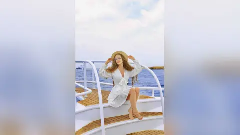 Smiling woman in a white summer dress and sun hat relaxing on a boat deck under a bright blue sky.