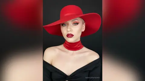 Glamorous woman in a black off-the-shoulder top and wide-brimmed red hat posing against a dark background.