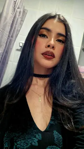 DashaRodriiguez poses with long dark hair, wearing a black choker and a dark floral low-cut top against a bathroom background.