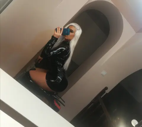 Glamorous woman in a black shiny latex outfit taking a mirror selfie in a modern home setting.