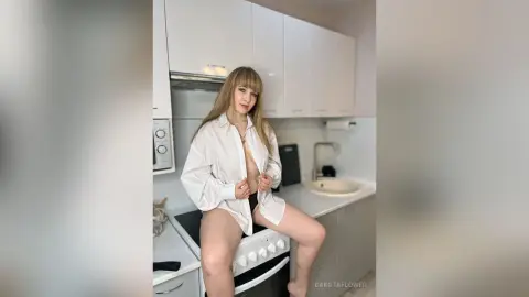 Blonde woman in an oversized white button-down shirt sitting on a kitchen stove in a bright modern home.