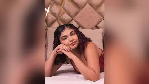 Young woman with wavy dark hair in a red lace camisole relaxing on a bed with a cozy, warm mood.