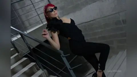 Stylish woman in black outfit and sunglasses posing on concrete stairs in a moody urban setting.