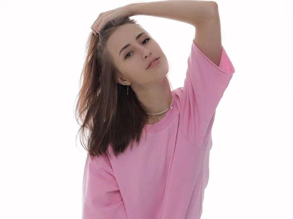 NoellaGlowinski poses with one hand in her long brown hair, wearing a pink t-shirt against a plain white background.