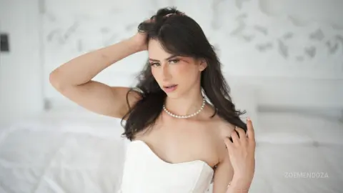 Elegant woman in a white strapless dress and pearl necklace posing in a bright, minimalist bedroom.
