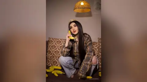 JuliDeming wearing a crown and a plaid shirt sits on a patterned couch, biting into a banana.