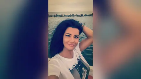 MissyJolie poses outdoors by the ocean wearing a white t-shirt with her hand in her dark hair.