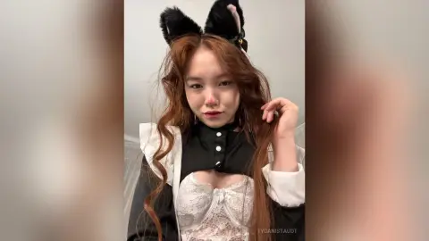 Young woman with long wavy hair wearing black cat ears and a white lace corset in a soft indoor setting.
