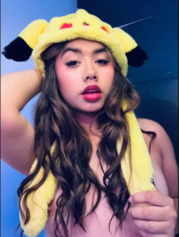 Young woman with long wavy hair wearing a playful yellow Pikachu plush hat in a soft, moody indoor setting.