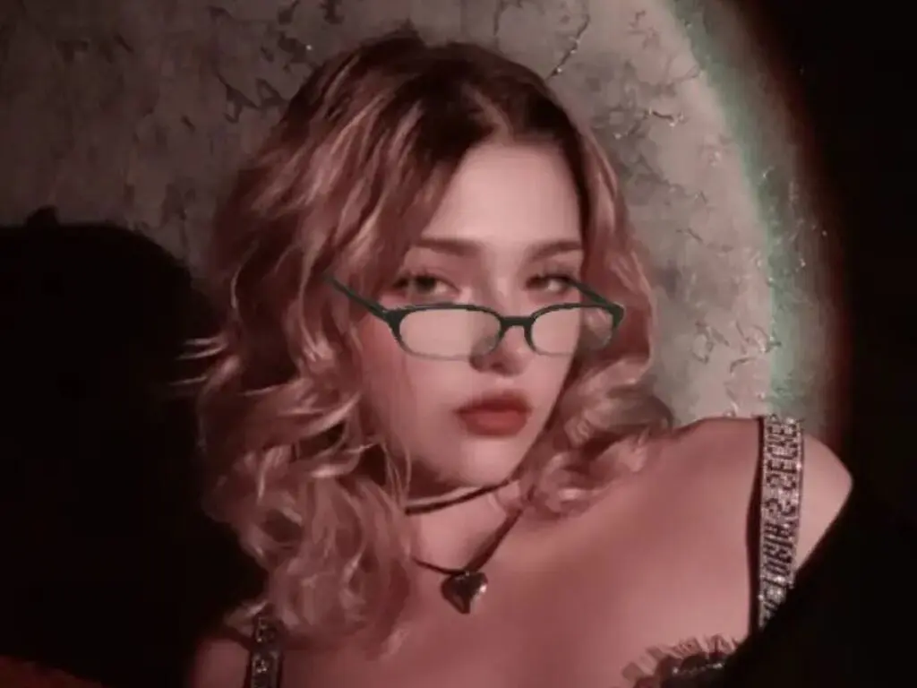 Blonde woman with green glasses and heart necklace posing in moody, low-light indoor setting.