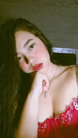 Young woman with long dark hair wearing a red lace top and silver necklace in soft indoor lighting.
