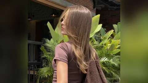 Side profile of a woman with long blonde hair wearing a brown t-shirt in a lush tropical garden setting.