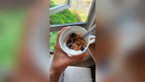 Hand holding a white bowl of healthy breakfast yogurt topped with fresh blueberries and granola by a window.