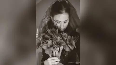 Melancholic bride in a dark lace wedding dress holding a large bouquet of flowers in a moody black and white portrait.