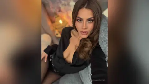 AnaMathers poses while wearing a low-cut black blouse, showing cleavage and her long brown hair against a grey chair.
