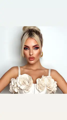 Glamorous blonde woman in a white floral corset top with elegant makeup against a plain background.