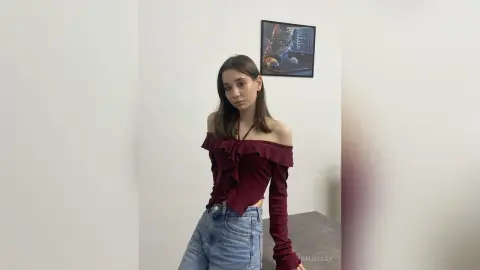 Young woman in a burgundy off-the-shoulder ruffle top and blue jeans posing in a minimalist white room.