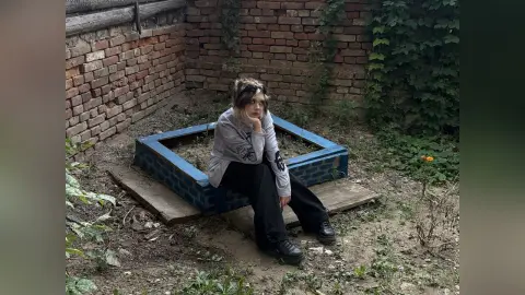 VernellZiccardi sits on a blue brick ledge outdoors, wearing a grey long-sleeve shirt and black pants with her hand resting on her chin.