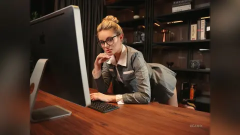BeaNix wears glasses and a grey button-down shirt while leaning over a wooden desk in front of a computer monitor.