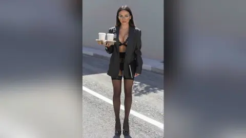 Stylish woman in a black blazer and sheer lingerie holding coffee cups on a sunny street.