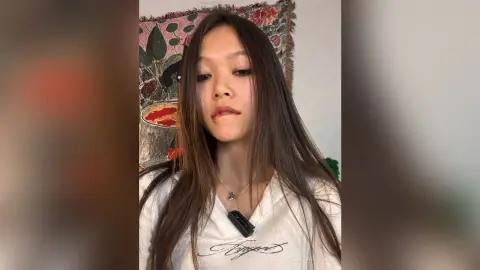 Young woman with long brown hair wearing a white graphic t-shirt in a cozy indoor setting.