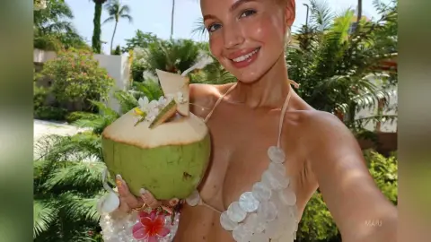 Smiling woman in a white shell bikini holding a fresh coconut drink at a tropical summer resort.
