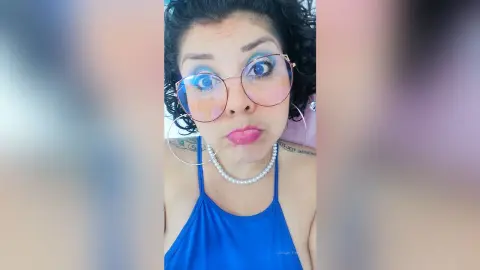 Smiling woman with curly hair and glasses wearing a blue halter top and pearl necklace in a bright setting.