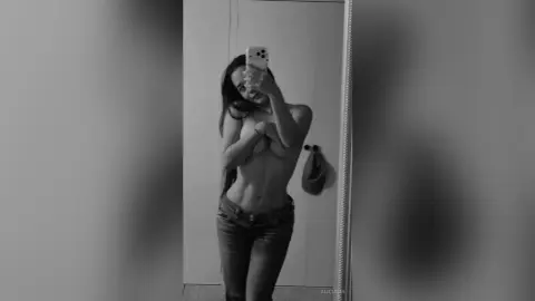 Black and white mirror selfie of a young woman in denim jeans posing in a minimalist room.