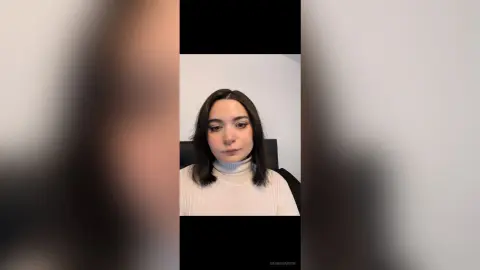 Young woman with dark hair wearing a white turtleneck sweater in a minimalist indoor setting.
