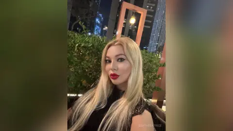 Blonde woman in a black lace outfit posing with glamorous makeup in a modern urban setting at night.