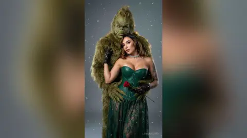 Woman in a green lace gown holding a red rose while being embraced by the Grinch in a snowy winter setting.