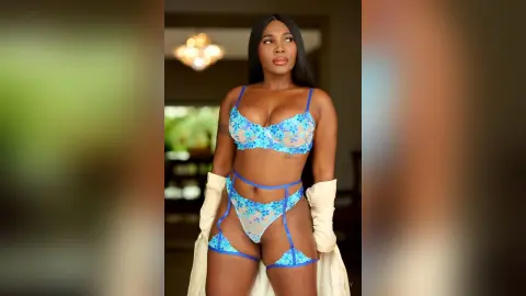 Confident Black woman in blue floral lace lingerie set posing in a warm, sunlit indoor setting.