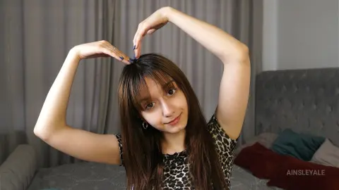 Young woman with long brown hair in a leopard print top making a heart shape with her hands in a cozy bedroom.