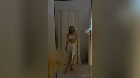 Young woman in a white floral cutout maxi dress taking a mirror selfie in a minimalist bedroom.