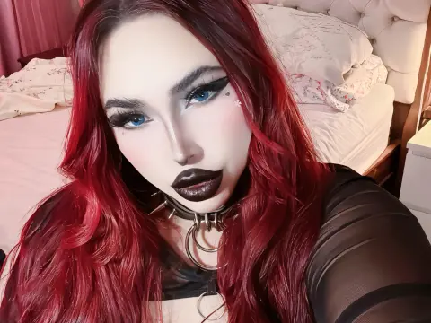 Woman with long red hair and dark gothic makeup wearing a black choker in a bedroom setting.