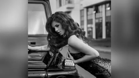 RubyAttwood poses against a black car, wearing a dark, shimmering patterned top while looking down with her long wavy hair falling over her shoulder.