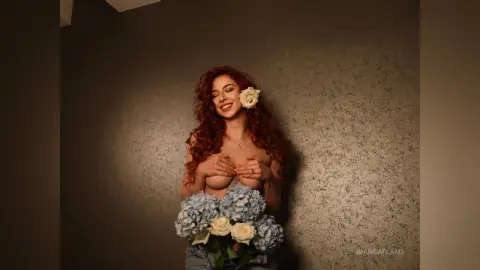 Smiling redhead woman with flowers holding a bouquet against a textured wall in a warm, moody setting.