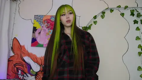 Young woman with long green hair wearing a plaid flannel shirt in a room with colorful anime wall art.