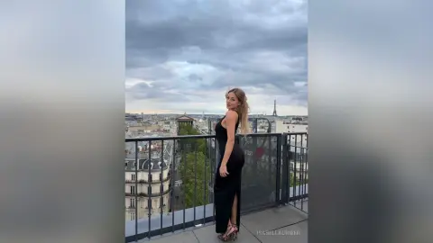 Elegant woman in a black evening gown posing on a Parisian balcony overlooking the Eiffel Tower under cloudy skies.