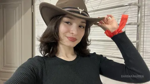 Smiling woman in a black ribbed sweater and brown cowboy hat posing in a bright indoor setting.