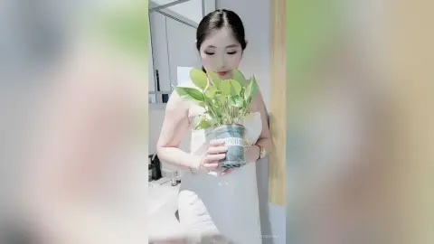 LouiseAnna holds a green leafy plant in a pot, wearing a white strapless top with her dark hair pulled back in a bathroom setting.