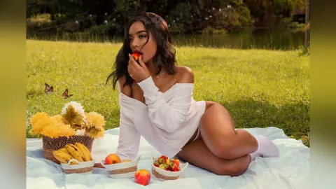 Young woman in a white shirt enjoying a summer picnic with fresh fruit on a sunny grass field.