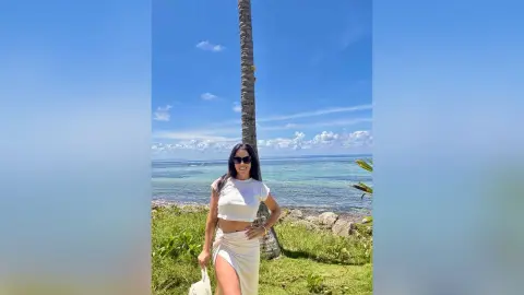 Smiling woman in white summer outfit and sunglasses standing by a palm tree on a sunny tropical beach.