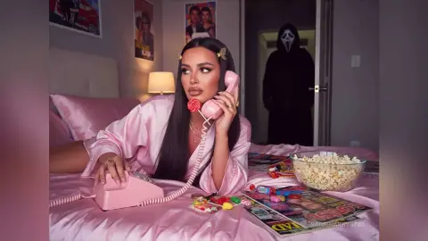 Woman in pink satin pajamas talking on a vintage pink phone in a bedroom with a Ghostface figure in the background.