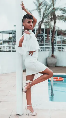 AnnPrice poses outdoors by a pool in a short white dress and white high heels, leaning against a white pole with one leg bent.